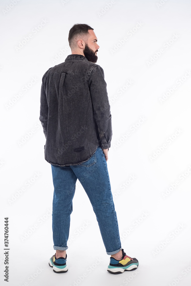 A bearded man in denim clothes stands with back. Full-length portrait of a man. Portrait of a man with a beard on a white background. The guy in jeans and sneakers. A bearded man in denim clothes stands with back. Full-length portrait of a man. Portrait of a man with a beard on a white background. The guy in jeans and sneakers.
