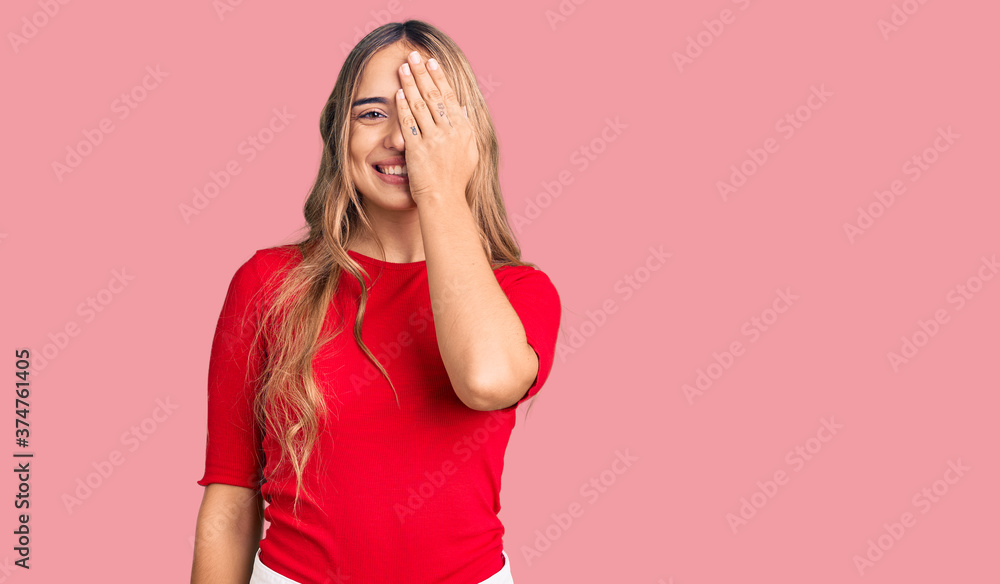 Young beautiful blonde woman wearing casual clothes covering one eye with hand, confident smile on face and surprise emotion. Young beautiful blonde woman wearing casual clothes covering one eye with hand, confident smile on face and surprise emotion.