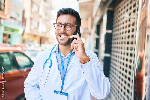 Young handsome hispanic doctor wearing stethoscope smiling happy Standing with smile on face talking on the smartphone at town street.