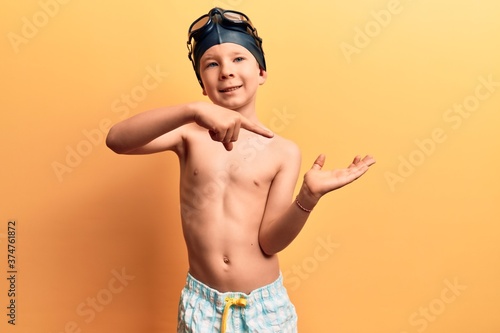 Cute blond kid wearing swimwear and swimmer glasses amazed and smiling to the camera while presenting with hand and pointing with finger. © Krakenimages.com