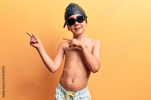 Cute blond kid wearing swimwear and swimmer glasses smiling and looking at the camera pointing with two hands and fingers to the side. © Krakenimages.com