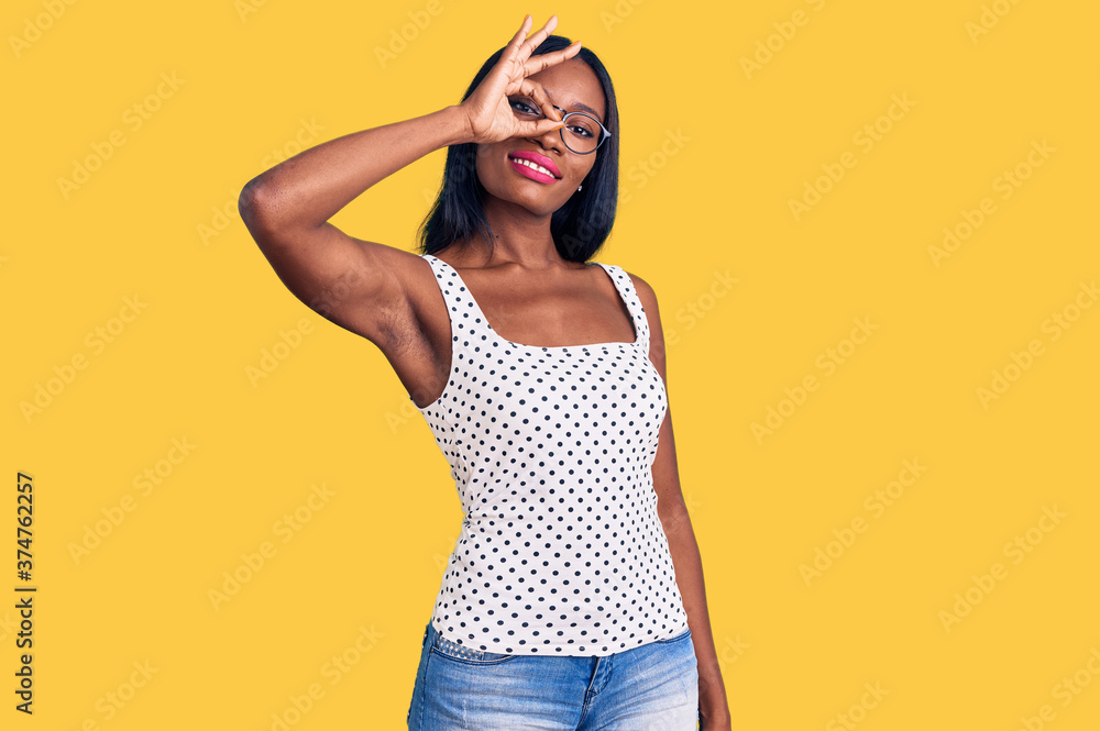 Young african american woman wearing casual clothes and glasses smiling happy doing ok sign with hand on eye looking through fingers Young african american woman wearing casual clothes and glasses smiling happy doing ok sign with hand on eye looking through fingers