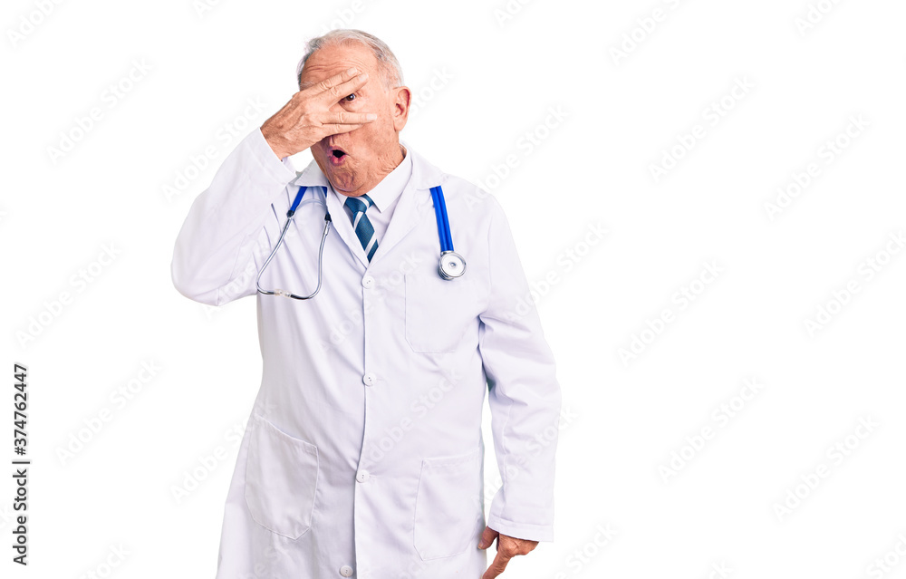 Senior handsome grey-haired man wearing doctor coat and stethoscope peeking in shock covering face and eyes with hand, looking through fingers with embarrassed expression. Senior handsome grey-haired man wearing doctor coat and stethoscope peeking in shock covering face and eyes with hand, looking through fingers with embarrassed expression.