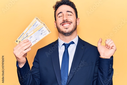 Young hispanic man wearing suit holding boarding pass screaming proud, celebrating victory and success very excited with raised arm