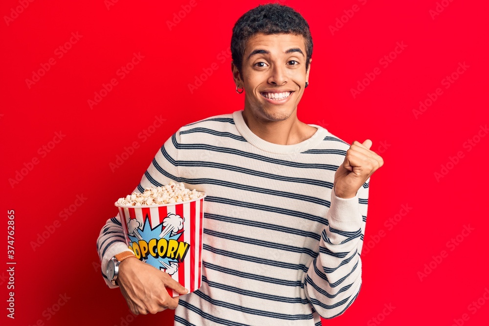 Fototapeta premium Young african amercian man holding popcorn screaming proud, celebrating victory and success very excited with raised arm