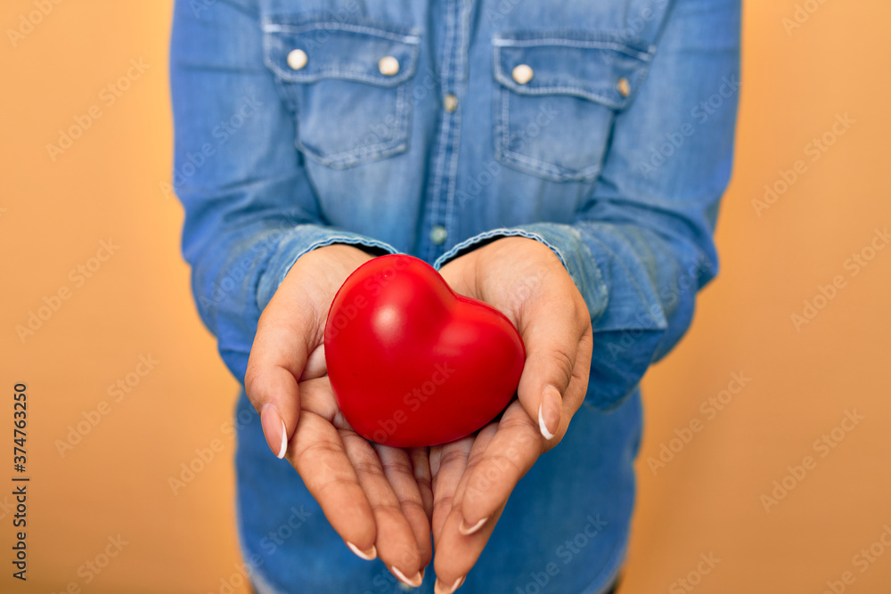 Woman holding red heart with two hands standing over isolated yellow background