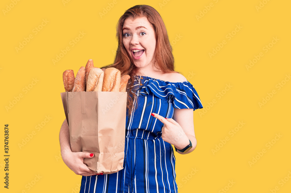 Obraz premium Young beautiful redhead woman holding paper bag with bread smiling happy pointing with hand and finger