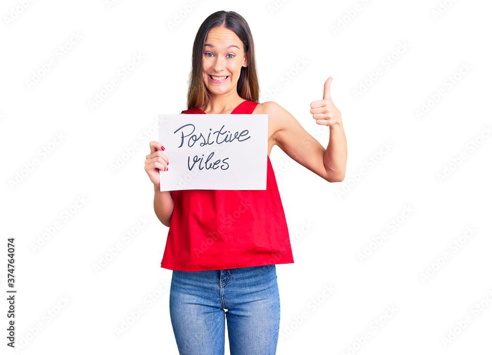 Beautiful brunette young woman holding positive vibes banner smiling happy and positive, thumb up doing excellent and approval sign Beautiful brunette young woman holding positive vibes banner smiling happy and positive, thumb up doing excellent and approval sign