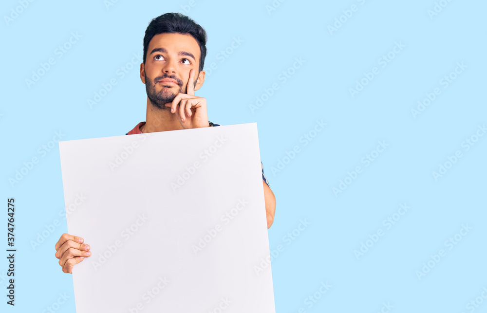 Young handsome hispanic man holding cardboard banner with blank space serious face thinking about question with hand on chin, thoughtful about confusing idea Young handsome hispanic man holding cardboard banner with blank space serious face thinking about question with hand on chin, thoughtful about confusing idea