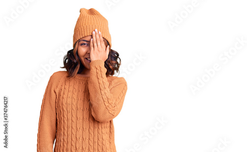 Young beautiful mixed race woman wearing wool sweater and winter hat covering one eye with hand, confident smile on face and surprise emotion. photo