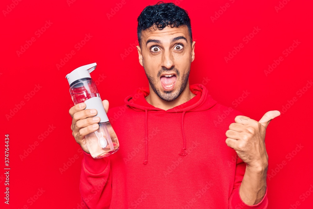 Young hispanic man drinking bottle of water pointing thumb up to the side smiling happy with open mouth Young hispanic man drinking bottle of water pointing thumb up to the side smiling happy with open mouth