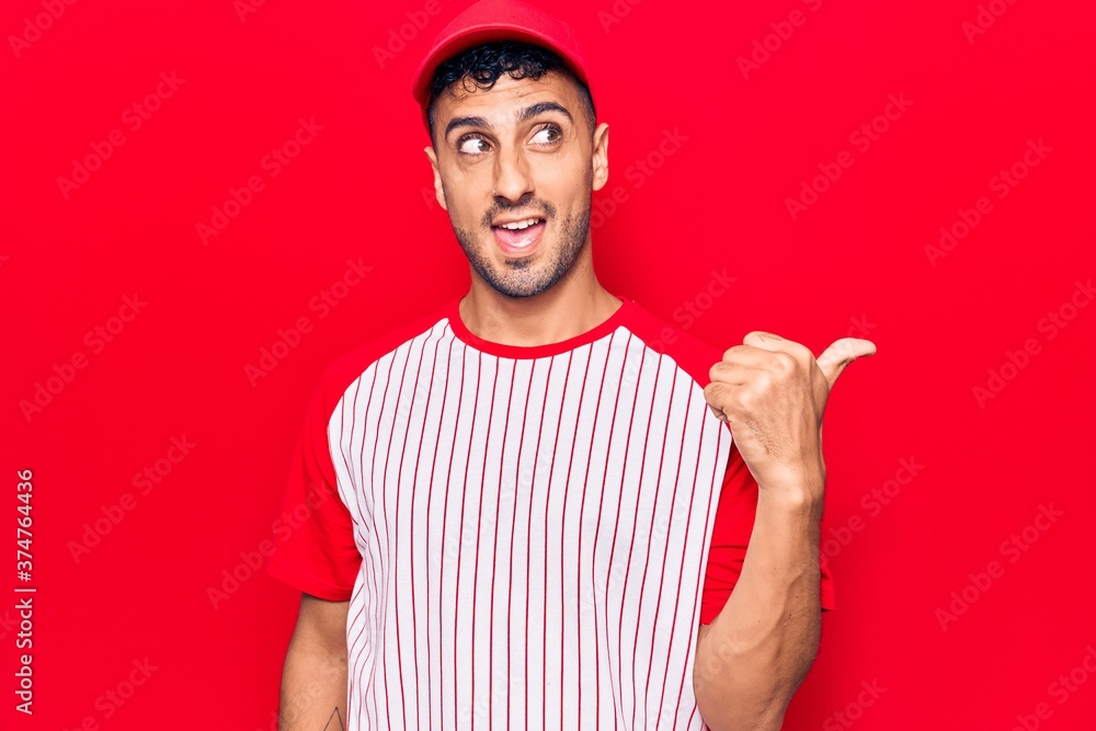 Young hispanic man wearing baseball uniform pointing thumb up to the side smiling happy with open mouth Young hispanic man wearing baseball uniform pointing thumb up to the side smiling happy with open mouth