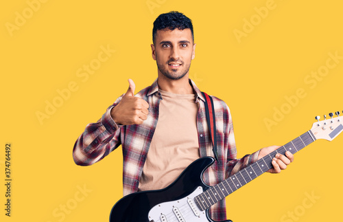 Young hispanic man playing electric guitar smiling happy and positive, thumb up doing excellent and approval sign