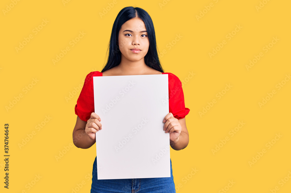 Young beautiful asian girl holding blank empty banner thinking attitude and sober expression looking self confident Young beautiful asian girl holding blank empty banner thinking attitude and sober expression looking self confident