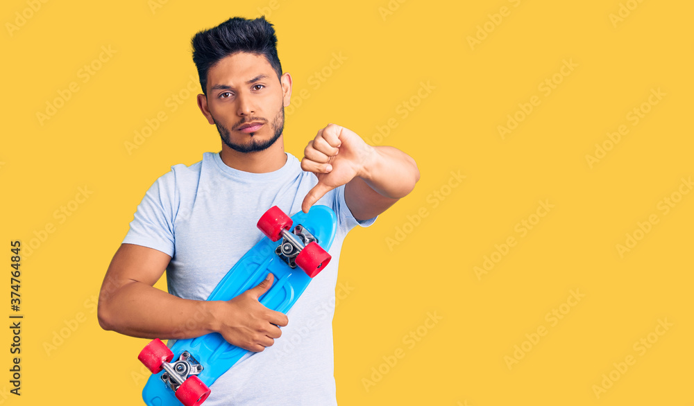 Handsome latin american young man holding skate with angry face, negative sign showing dislike with thumbs down, rejection concept Handsome latin american young man holding skate with angry face, negative sign showing dislike with thumbs down, rejection concept