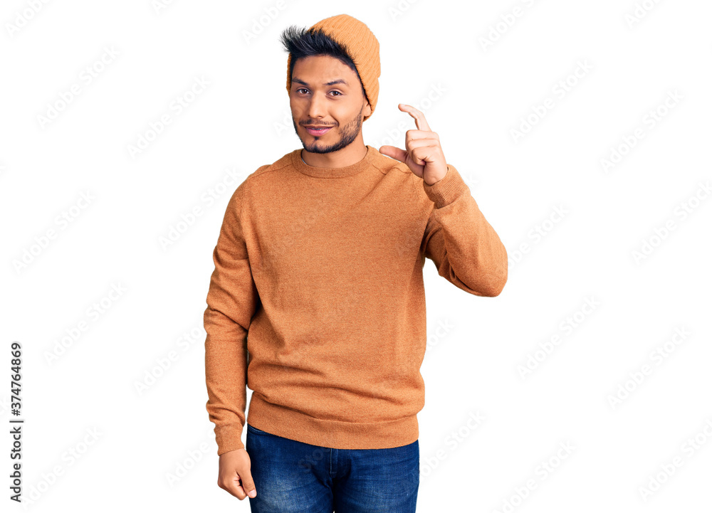 Handsome latin american young man wearing winter sweater and wool hat smiling and confident gesturing with hand doing small size sign with fingers looking and the camera. measure concept. Handsome latin american young man wearing winter sweater and wool hat smiling and confident gesturing with hand doing small size sign with fingers looking and the camera. measure concept.