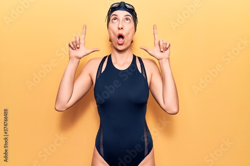Young beautiful hispanic woman wearing swimwear and swimmer glasses amazed and surprised looking up and pointing with fingers and raised arms.