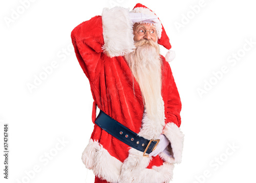 Old senior man with grey hair and long beard wearing traditional santa claus costume very happy and smiling looking far away with hand over head. searching concept.