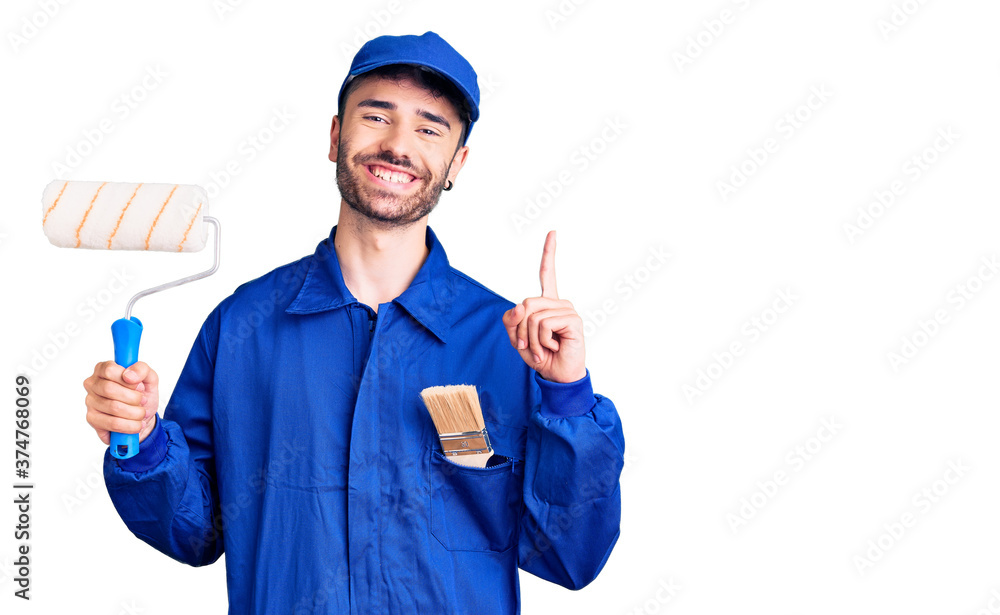 Young hispanic man wearing painter uniform holding roller surprised with an idea or question pointing finger with happy face, number one Young hispanic man wearing painter uniform holding roller surprised with an idea or question pointing finger with happy face, number one