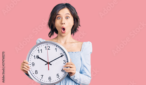 Young beautiful girl holding big clock scared and amazed with open mouth for surprise, disbelief face