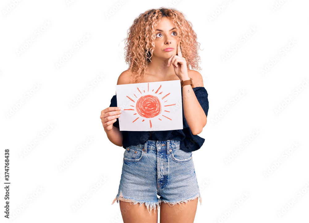 Young blonde woman with curly hair holding sun draw serious face thinking about question with hand on chin, thoughtful about confusing idea Young blonde woman with curly hair holding sun draw serious face thinking about question with hand on chin, thoughtful about confusing idea