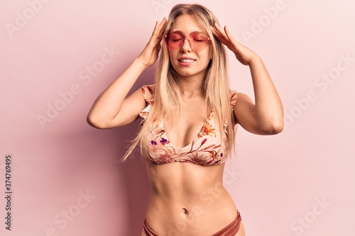 Young beautiful blonde woman wearing bikini heart sunglasses with hand on head, headache because stress. suffering migraine.