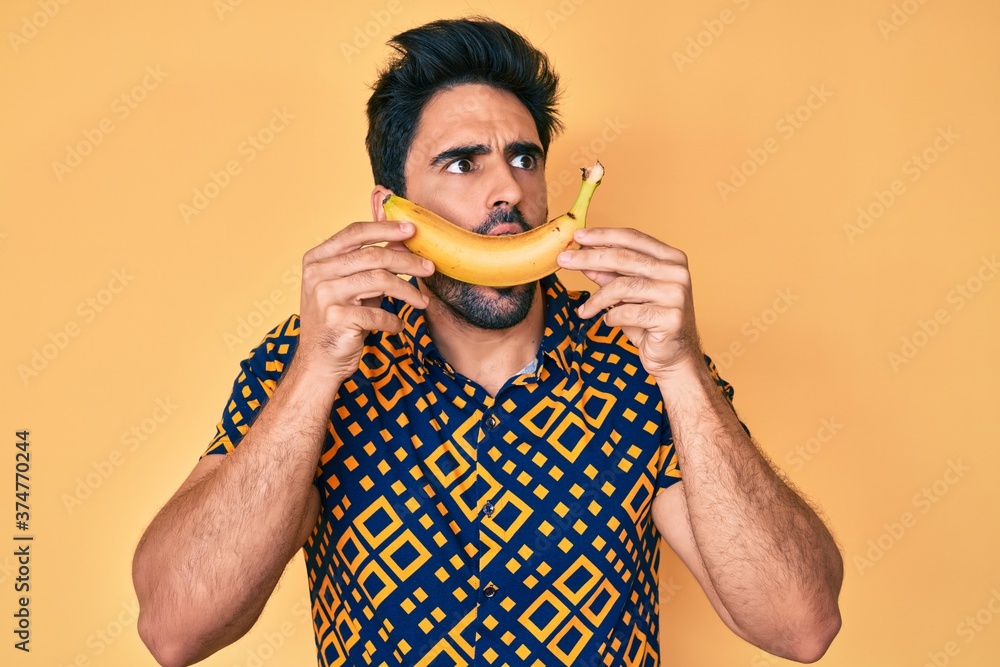 Handsome hispanic man with beard holding banana like funny smile angry and mad screaming frustrated and furious, shouting with anger. rage and aggressive concept. Handsome hispanic man with beard holding banana like funny smile angry and mad screaming frustrated and furious, shouting with anger. rage and aggressive concept.