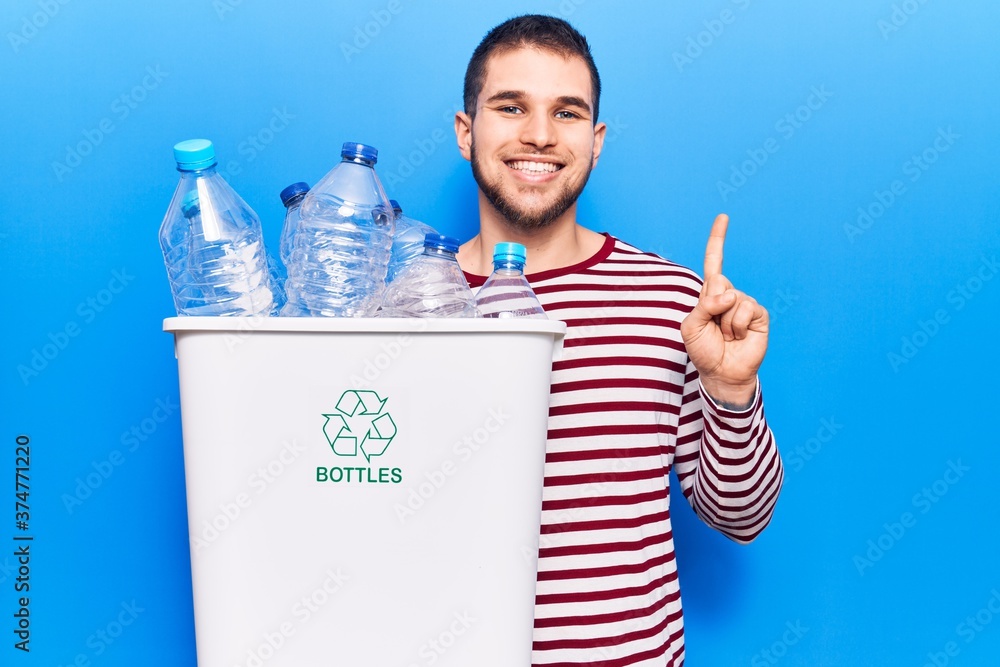 Young handsome man recycling plastic bottles smiling with an idea or question pointing finger with happy face, number one Young handsome man recycling plastic bottles smiling with an idea or question pointing finger with happy face, number one