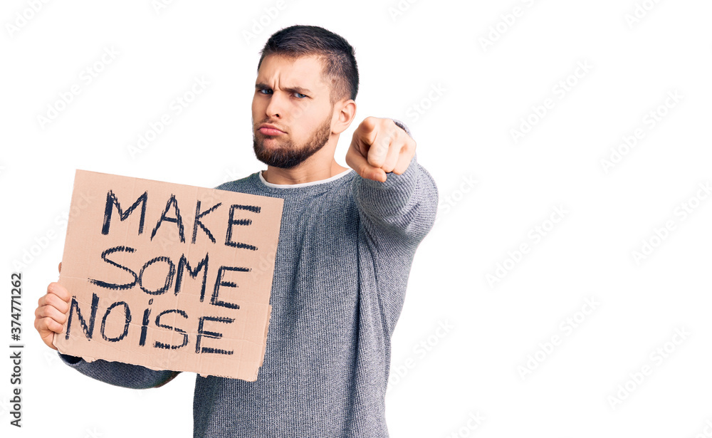 Young handsome man holding make some noise banner pointing with finger to the camera and to you, confident gesture looking serious Young handsome man holding make some noise banner pointing with finger to the camera and to you, confident gesture looking serious