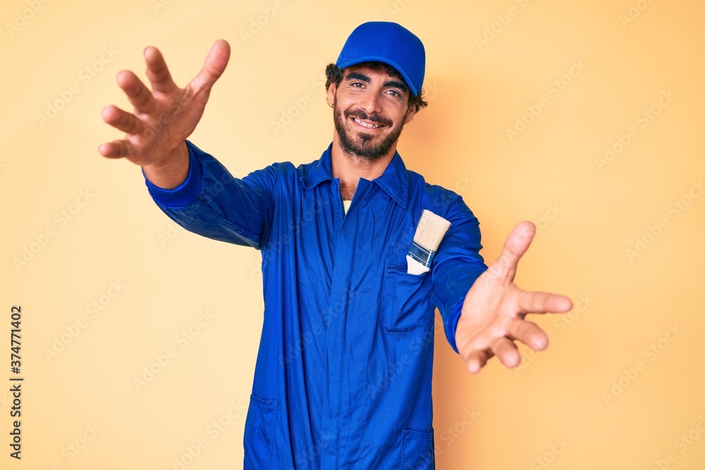 Handsome young man with curly hair and bear wearing builder jumpsuit uniform looking at the camera smiling with open arms for hug. cheerful expression embracing happiness. Handsome young man with curly hair and bear wearing builder jumpsuit uniform looking at the camera smiling with open arms for hug. cheerful expression embracing happiness.