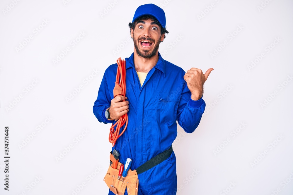 Handsome young man with curly hair and bear wearing builder jumpsuit uniform and electric cables pointing thumb up to the side smiling happy with open mouth