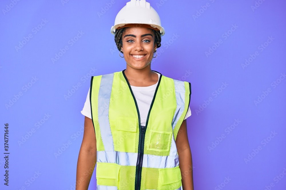 Young african american woman with braids wearing safety helmet and reflective jacket looking positive and happy standing and smiling with a confident smile showing teeth Young african american woman with braids wearing safety helmet and reflective jacket looking positive and happy standing and smiling with a confident smile showing teeth
