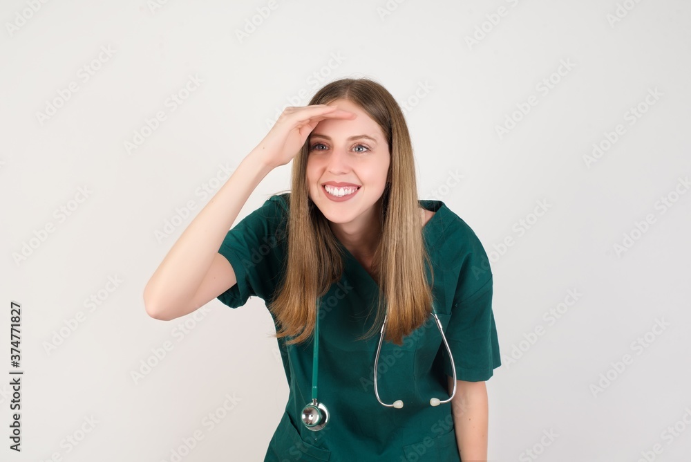 Female doctor wearing a green scrubs and stethoscope very happy and smiling looking far away with hand over head. Searching concept. Female doctor wearing a green scrubs and stethoscope very happy and smiling looking far away with hand over head. Searching concept.
