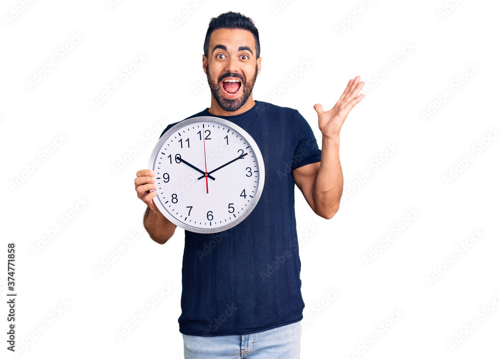 Young hispanic man holding big clock celebrating victory with happy smile and winner expression with raised hands Young hispanic man holding big clock celebrating victory with happy smile and winner expression with raised hands