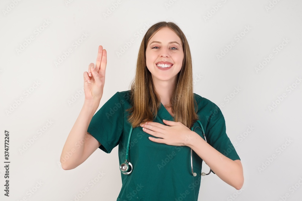 Female doctor wearing a green scrubs and stethoscope smiling swearing with hand on chest and fingers up, making a loyalty promise oath. Female doctor wearing a green scrubs and stethoscope smiling swearing with hand on chest and fingers up, making a loyalty promise oath.
