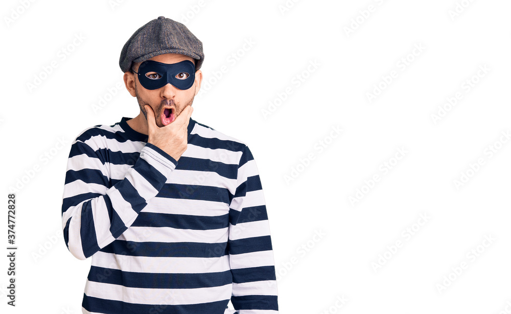 Fototapeta premium Young handsome man wearing burglar mask looking fascinated with disbelief, surprise and amazed expression with hands on chin