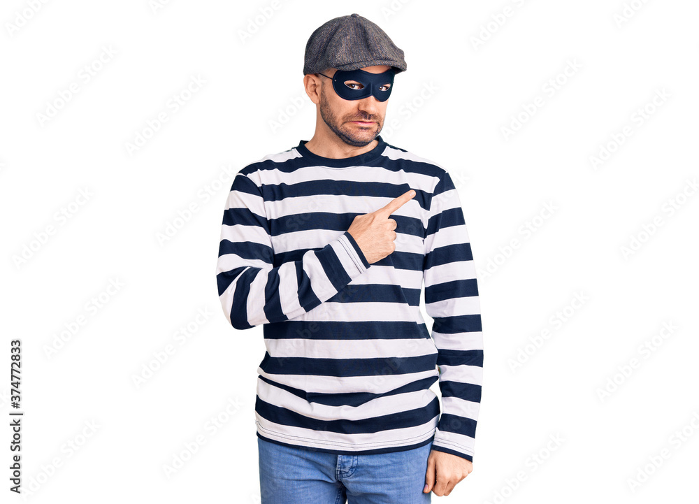 Young handsome man wearing burglar mask pointing aside worried and nervous with forefinger, concerned and surprised expression Young handsome man wearing burglar mask pointing aside worried and nervous with forefinger, concerned and surprised expression