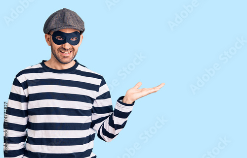 Young handsome man wearing burglar mask smiling cheerful presenting and pointing with palm of hand looking at the camera. © Krakenimages.com