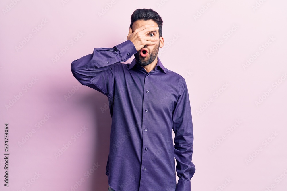 Young handsome man with beard wearing casual shirt peeking in shock covering face and eyes with hand, looking through fingers with embarrassed expression. Young handsome man with beard wearing casual shirt peeking in shock covering face and eyes with hand, looking through fingers with embarrassed expression.