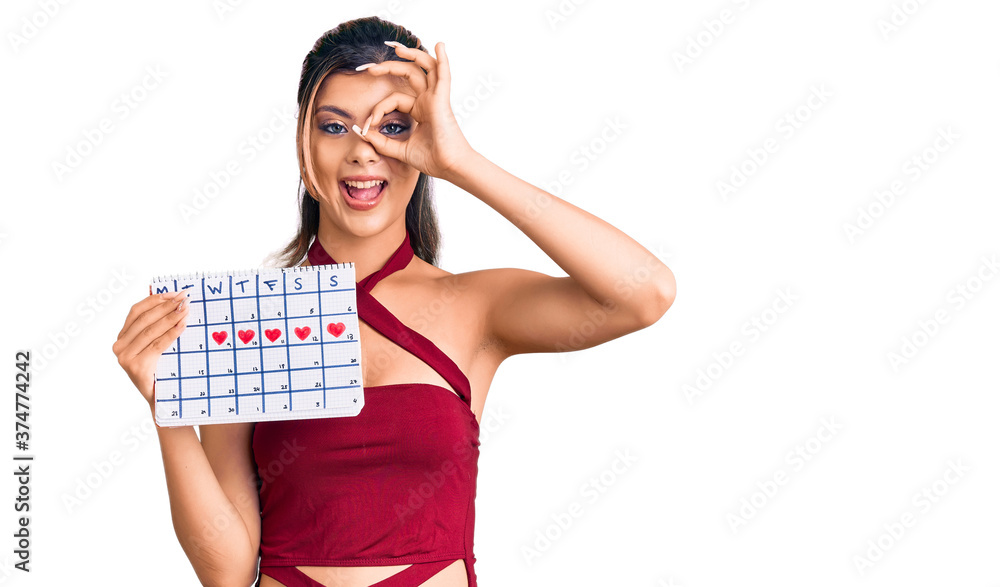 Young beautiful woman holding heart calendar smiling happy doing ok sign with hand on eye looking through fingers Young beautiful woman holding heart calendar smiling happy doing ok sign with hand on eye looking through fingers