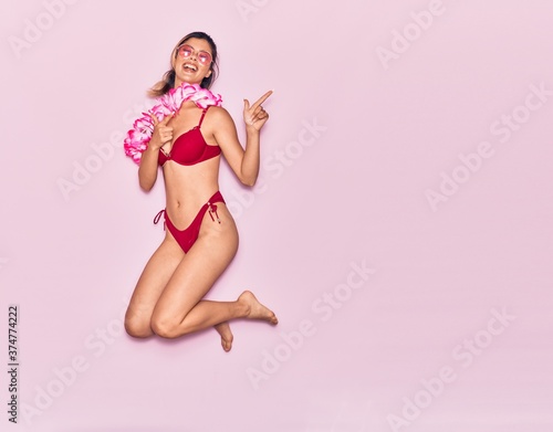 Young beautiful girl on vacation wearing bikini and hawaiian lei smiling happy. Jumping with smile on face pointing with fingers to the side over isolated pink background