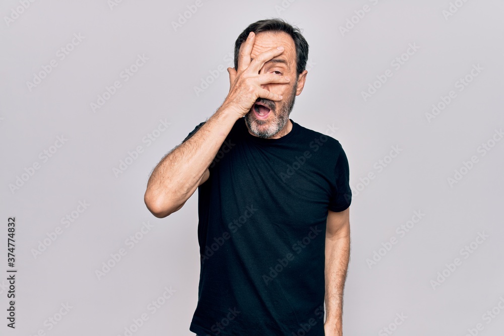 Middle age man wearing casual black t-shirt standing over isolated white background peeking in shock covering face and eyes with hand, looking through fingers afraid Middle age man wearing casual black t-shirt standing over isolated white background peeking in shock covering face and eyes with hand, looking through fingers afraid