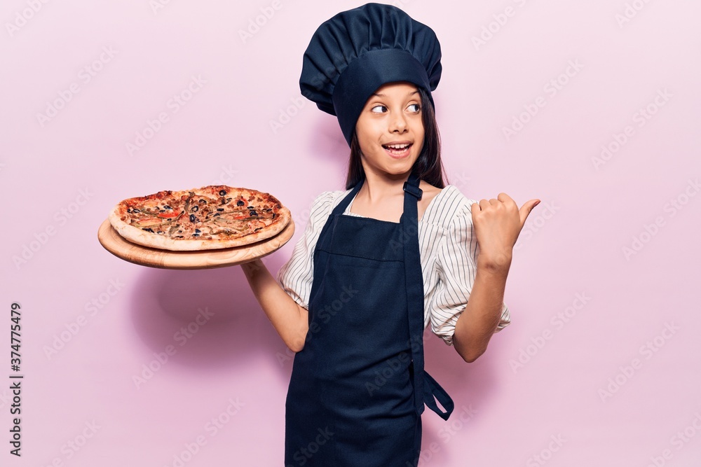 Beautiful child girl wearing cooker uniform holding italian pizza pointing thumb up to the side smiling happy with open mouth Beautiful child girl wearing cooker uniform holding italian pizza pointing thumb up to the side smiling happy with open mouth