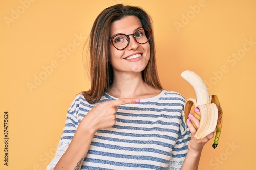 Beautiful caucasian woman eating banana as healthy snack smiling happy pointing with hand and finger