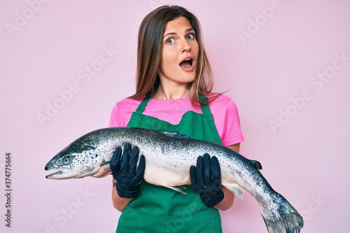 Beautiful caucasian woman fishmonger selling fresh raw salmon in shock face, looking skeptical and sarcastic, surprised with open mouth