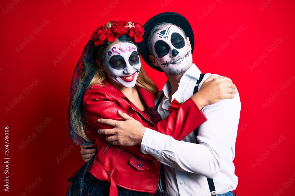 Couple wearing day of the dead costume over red hugging oneself happy and positive, smiling confident. self love and self care