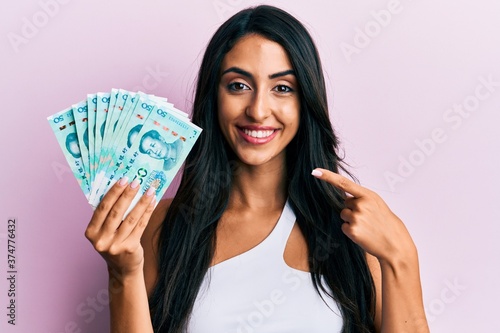 Beautiful hispanic woman holding yuan chinese banknotes smiling happy pointing with hand and finger