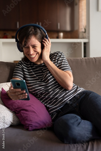 laughing woman sitting on a sofa with a smartphone in hand and headphones