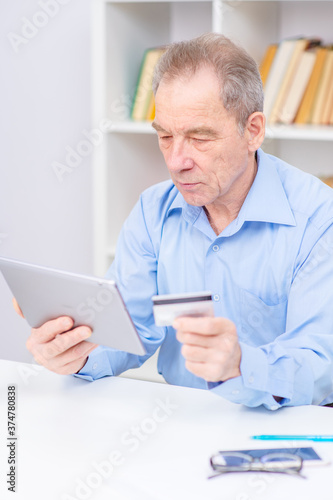 An elderly retired man makes online purchases using a tablet computer and a credit card. Shopping during lockdown concept