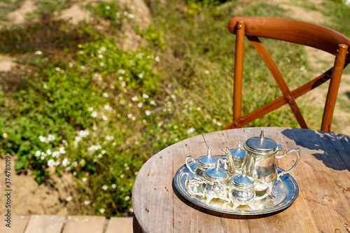 Silver tea pot set on tray are arranging on wooden vintage circle table with chair in the garden.Soft blured white flower on green grass background next to terrence in backyard.Afternoon tea concept. photo